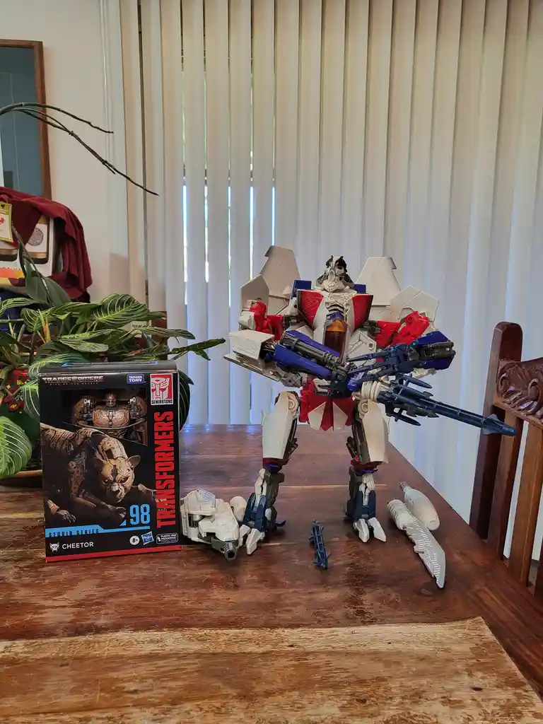 A large, white, red, and blue Transformers action figure stands on a wooden table next to its packaging, which is labeled…