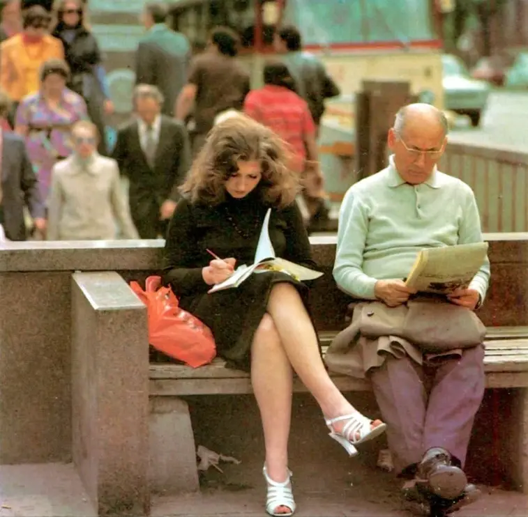 A woman in a black dress and white heels sits on a bench writing in a notebook, next to an older man in a light green…