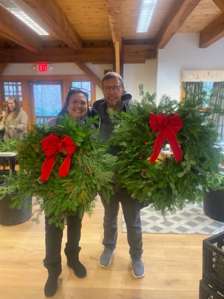 A smiling man and woman stand indoors holding large, identical Christmas wreaths decorated with red bows. They are in a room…