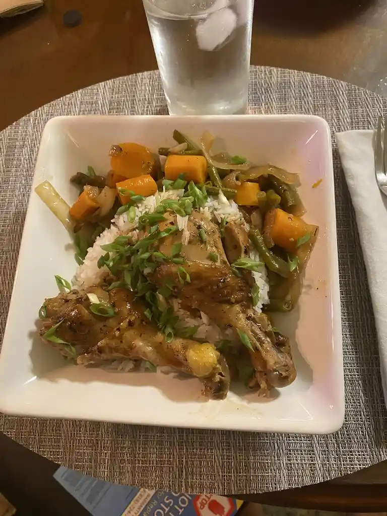 A close-up photo of a plated meal featuring two glazed chicken legs over white rice, topped with chopped green onions and…