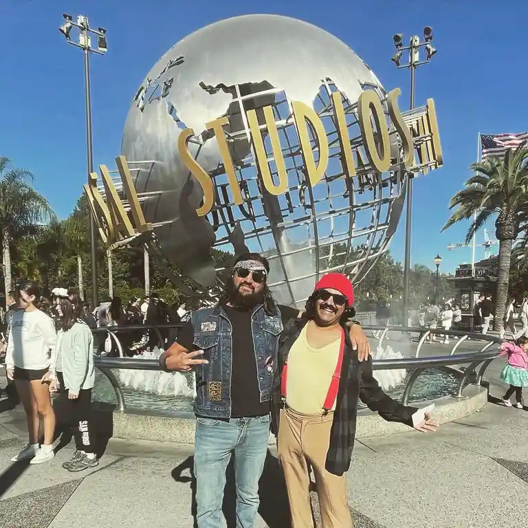 Two men posing in front of the iconic Universal Studios globe landmark, one dressed in a denim vest and bandana, the other…