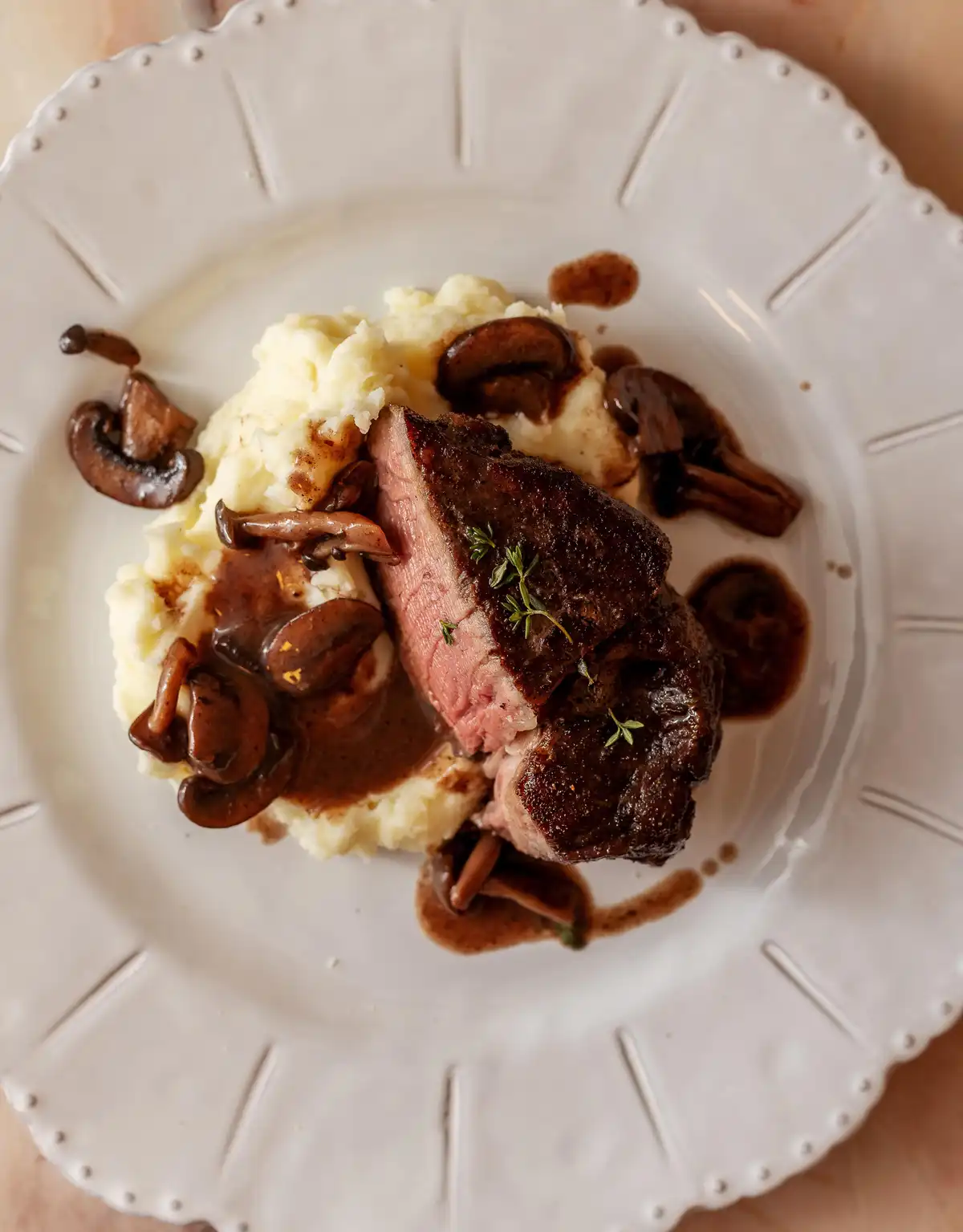 A top-down photo of a plated meal featuring a seared steak with a pink center, served atop creamy mashed potatoes and…