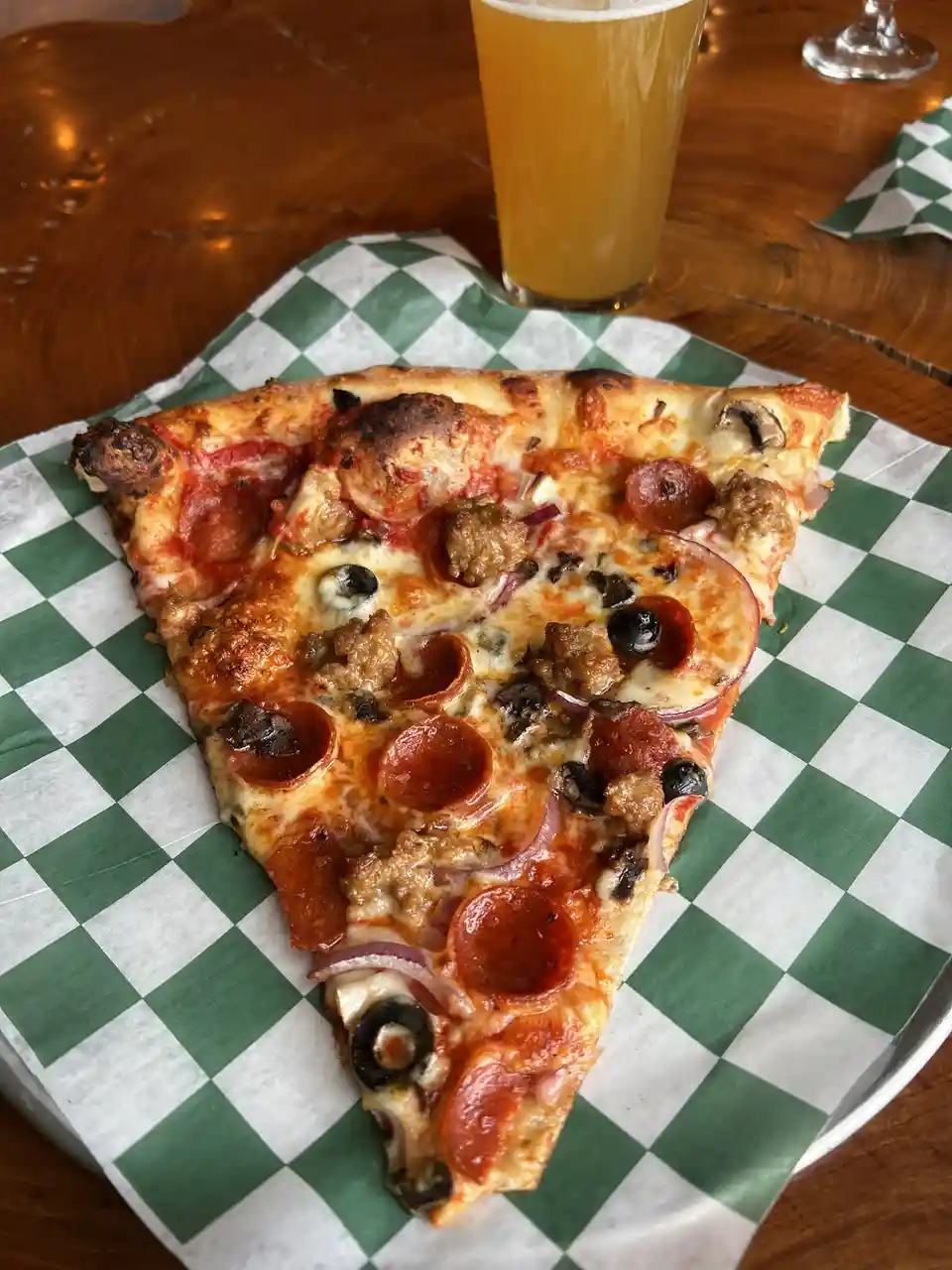 A single slice of loaded pizza with pepperoni, sausage, olives, and onions sits on green-and-white checkered paper on a…