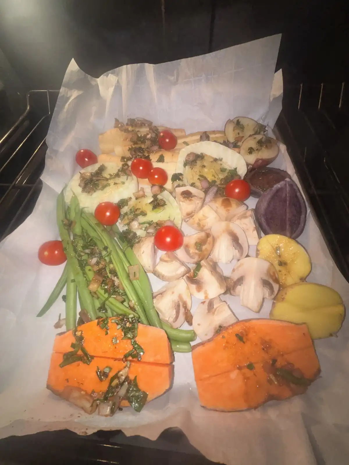 A baking tray lined with parchment paper holds a colorful assortment of roasted vegetables including sweet potato chunks…