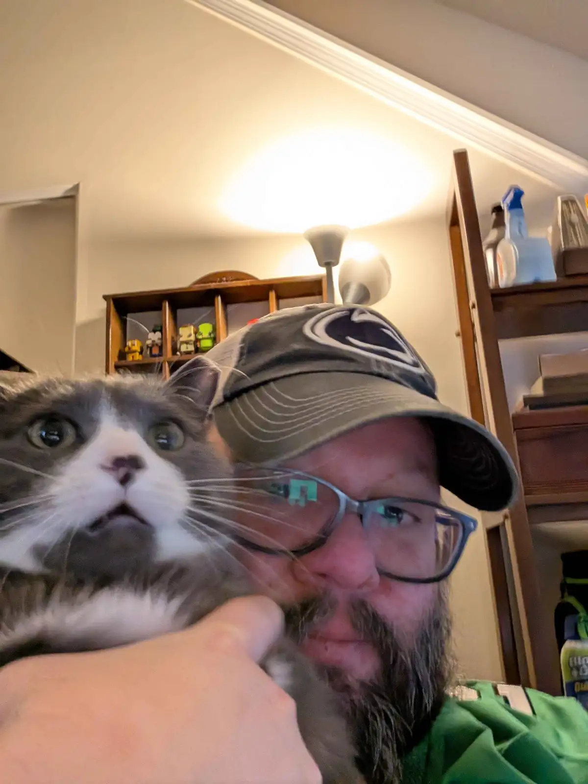 A man with a beard, glasses, and a baseball cap holds a gray and white cat close to the camera. The cat has wide, startled…