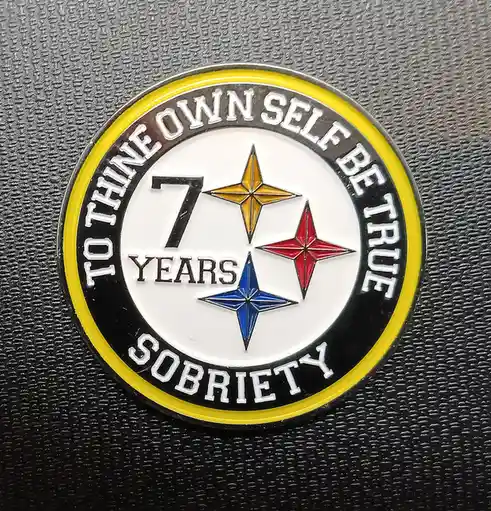 A close-up of a circular, metallic-looking sobriety chip with a black and yellow border, resting on a textured gray surface…