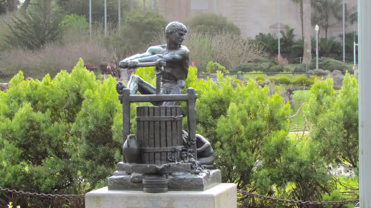 A bronze statue of a muscular man sitting atop a wine press, surrounded by green bushes, with overlaid text reading 'i'm not…