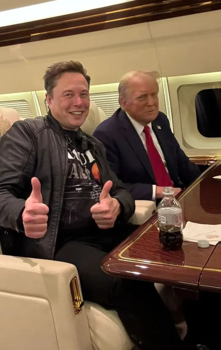 Two men sit side by side in a luxurious private jet cabin. The man on the left, wearing a black leather jacket over a…