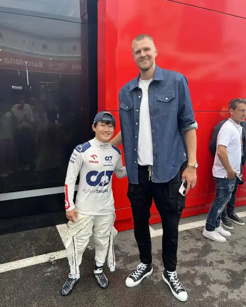A tall man in a denim shirt and black pants stands next to a much shorter man in a white racing suit with 'ALPHATAURI' and…