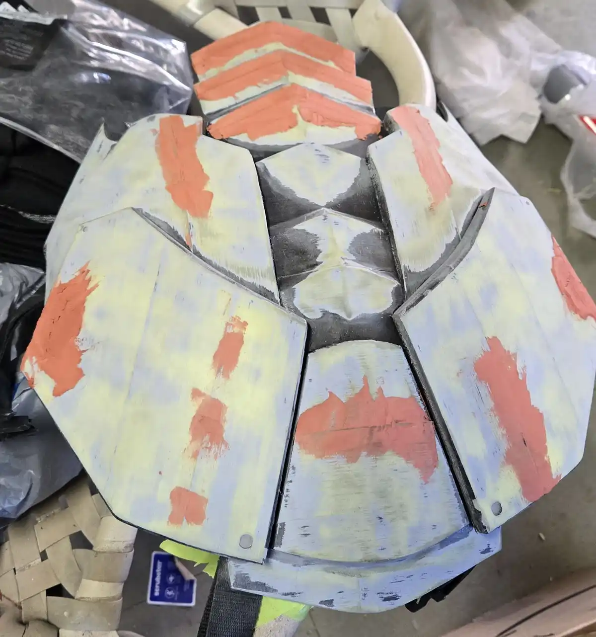 A close-up photo shows a partially painted, segmented armor piece resting on a chair, with uneven patches of orange, gray…