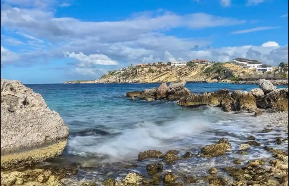 A scenic photo of a rocky coastline with turquoise water and waves crashing against rocks, with upscale houses visible on a…