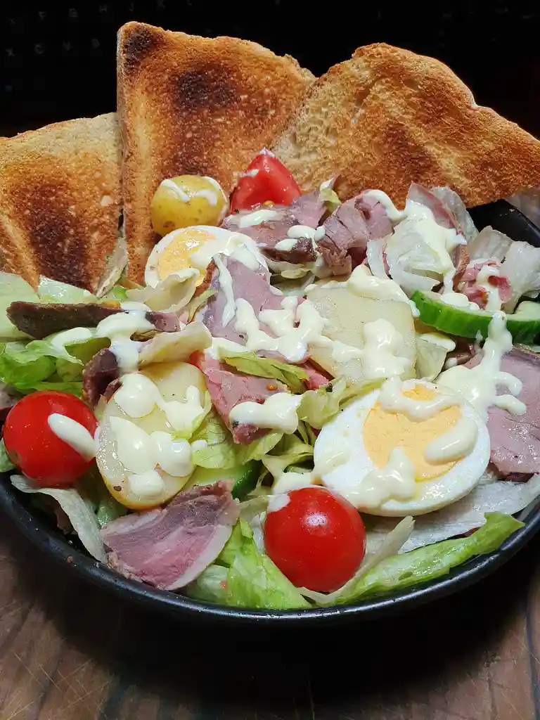 A close-up photo of a hearty, loaded salad in a black bowl, featuring lettuce, sliced boiled eggs, chunks of meat, cherry…