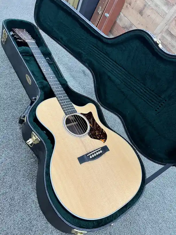 An acoustic guitar with a natural wood finish and cutaway body rests inside an open, black hardshell case lined with dark…