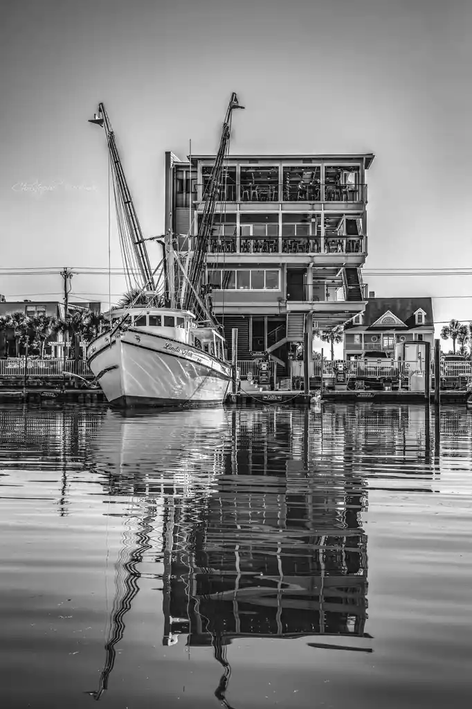 A black-and-white photo shows a shrimp boat named 'Linda Ann' docked beside a multi-story house built directly on pilings…