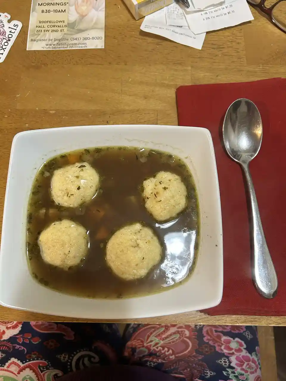 A white square bowl of broth with four round, pale dumplings floating in it sits on a wooden table next to a red napkin and…