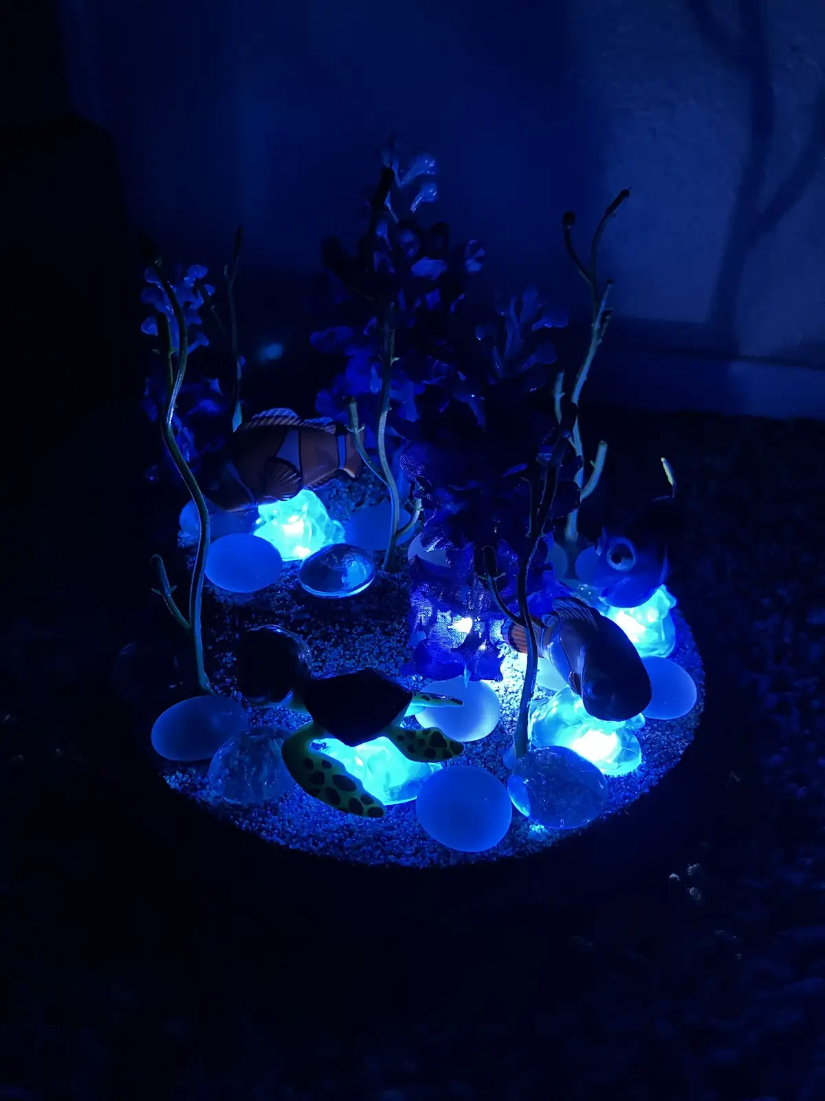 A dimly lit, circular decorative aquarium setup with glowing blue lights, featuring plastic fish and coral decorations, sits…
