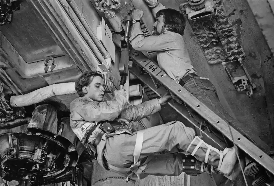 A black-and-white photo shows two men working in a cramped, industrial setting with pipes and machinery. One man is…