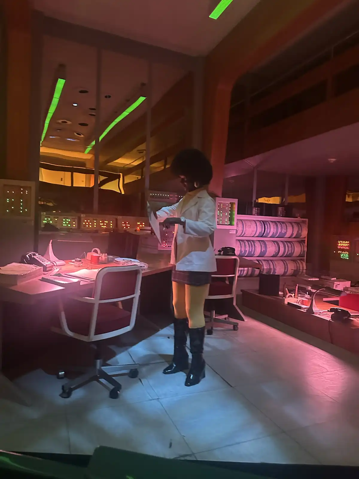 A person with a large afro, wearing a white lab coat, yellow tights, and black boots, stands in a retro-futuristic control…