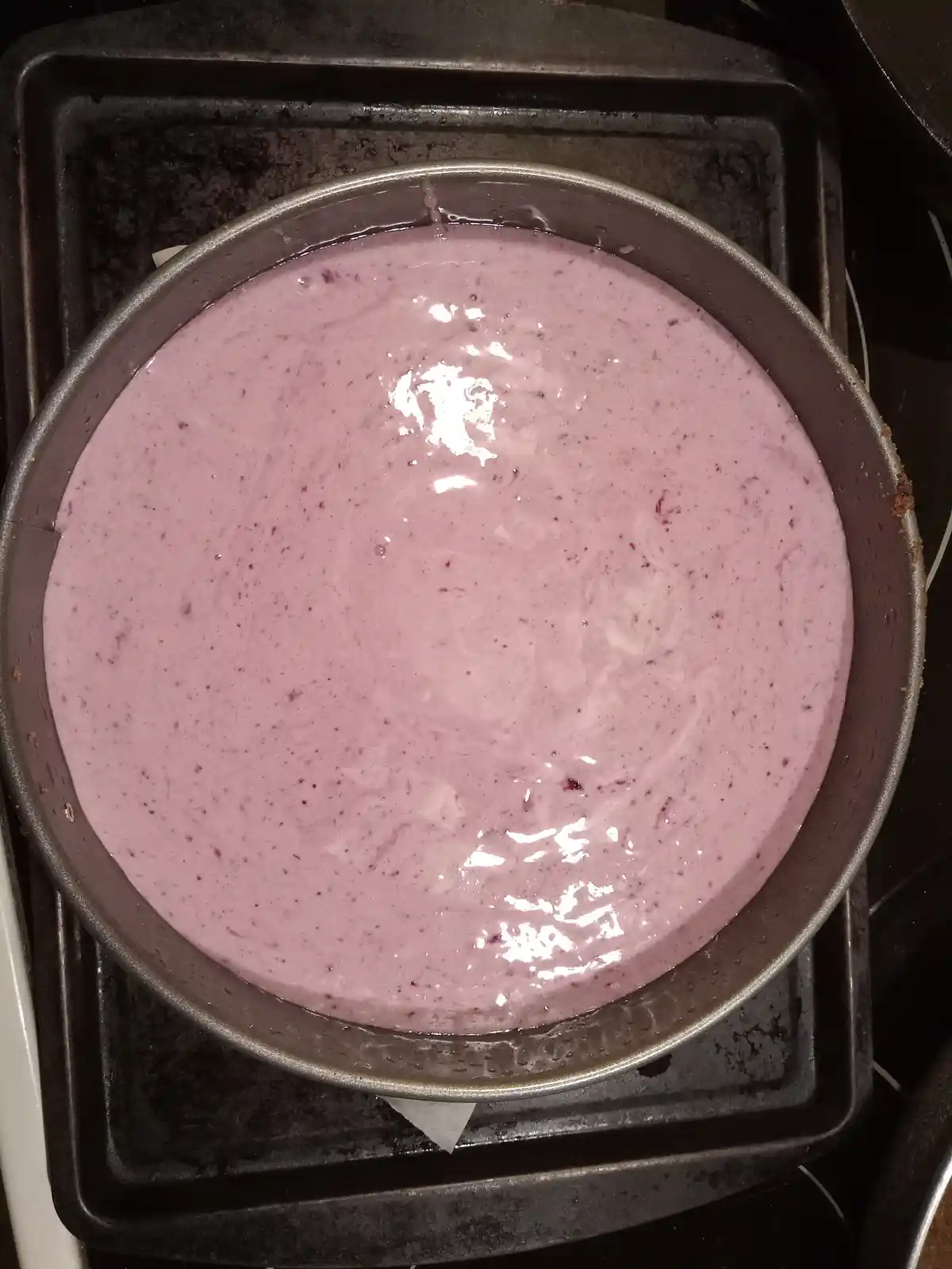 A top-down photo shows a round, light purple cake batter with small dark specks, sitting in a metal springform pan inside a…