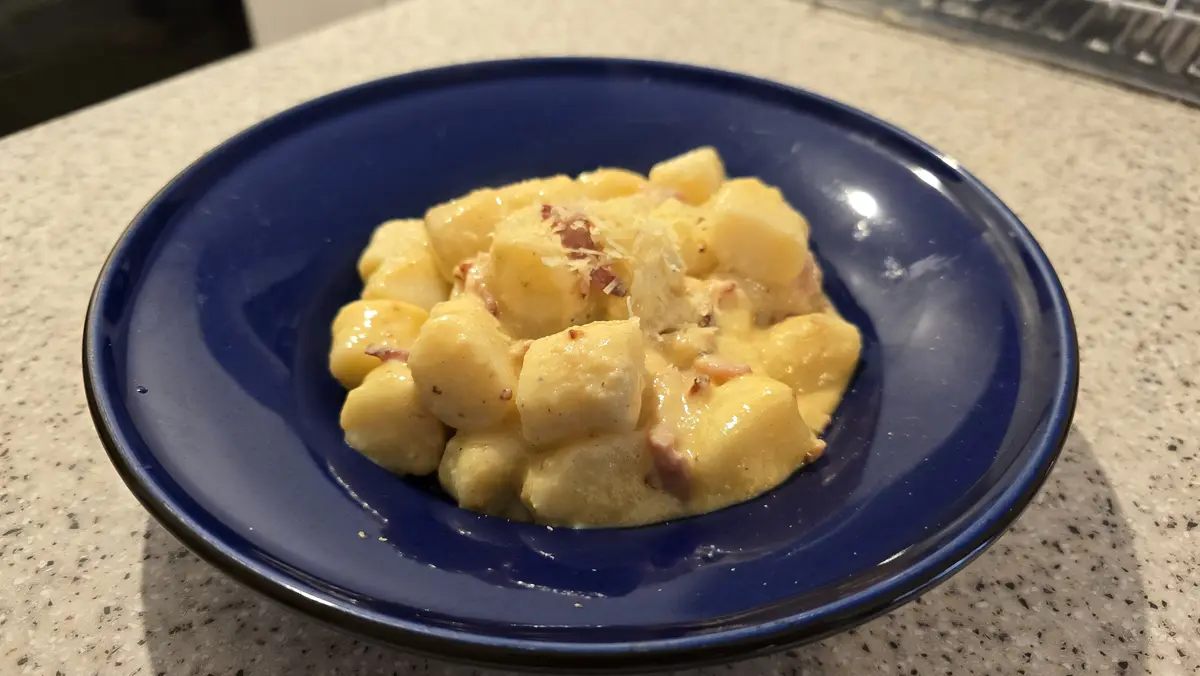 A close-up photo shows a blue ceramic bowl filled with creamy gnocchi, garnished with grated cheese and small pieces of…