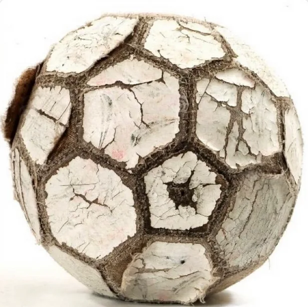 a close-up photograph of a heavily deteriorated soccer ball with cracked white panels and frayed brown stitching, set…