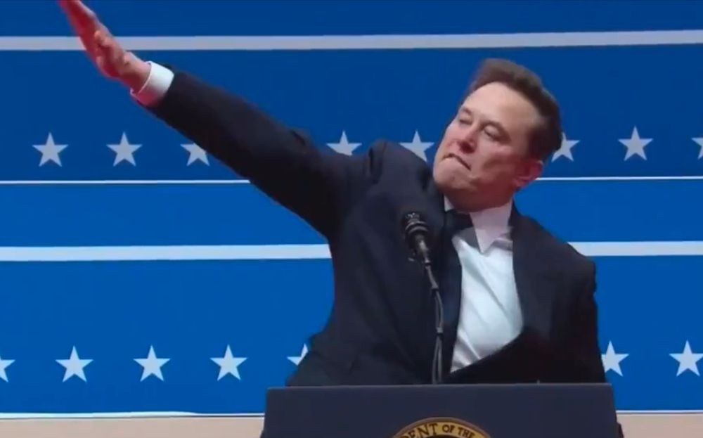 elon musk stands at a podium with a blue background featuring white stars and stripes, raising his right arm in a gesture…