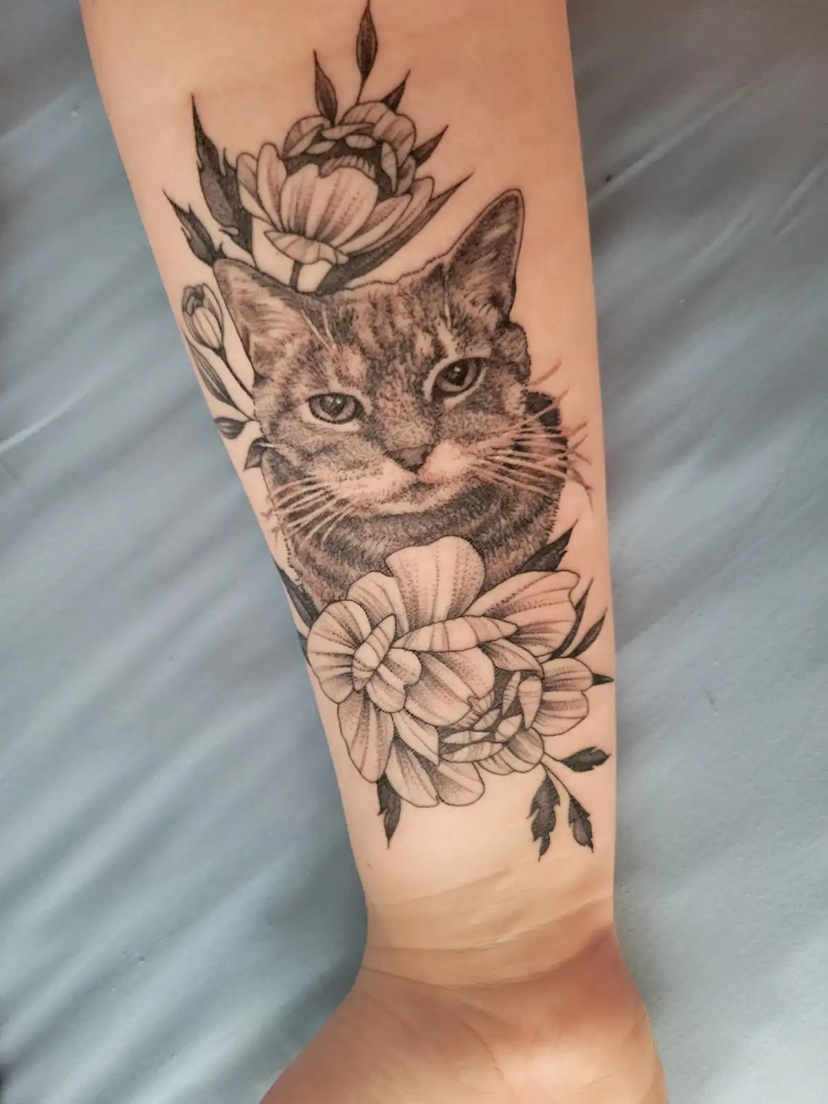 A black and grey tattoo on a person's forearm depicts a detailed cat's face surrounded by large, stylized flowers. The cat's…