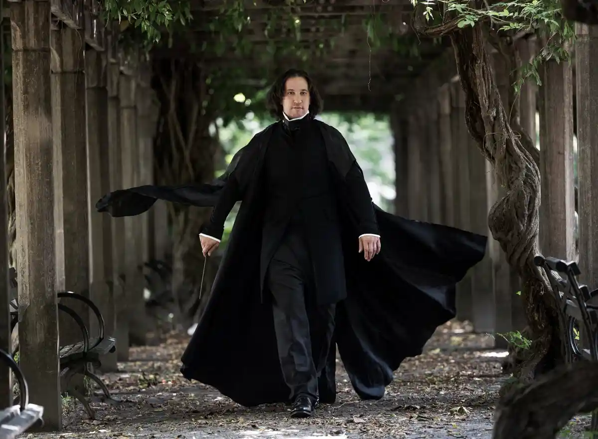 A man dressed as Severus Snape from Harry Potter walks confidently down a leafy, vine-covered wooden pergola, his long black…