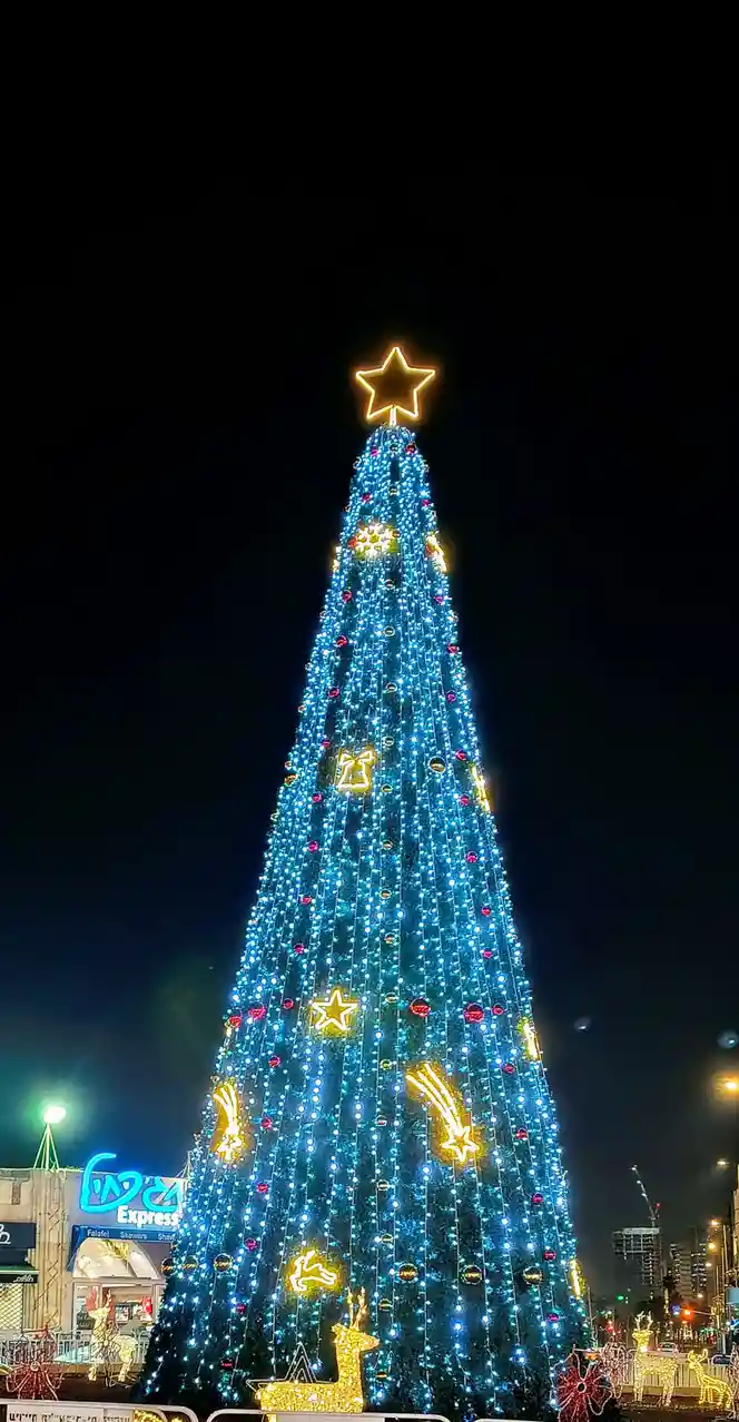 A tall, brightly lit Christmas tree adorned with blue lights, red ornaments, and golden star and reindeer decorations…