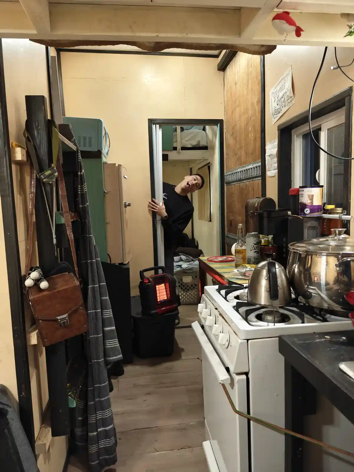 A man in a black shirt leans out of a doorway in a cluttered, cramped kitchen, laughing maniacally while holding the door…