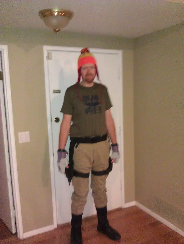 A man stands indoors in front of a white door, wearing a bright orange and yellow beanie with red tassels, a green t-shirt…