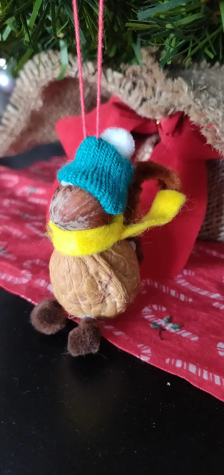 A handmade Christmas ornament shaped like a walnut, decorated with a blue knitted hat, a yellow scarf, and brown felt limbs…
