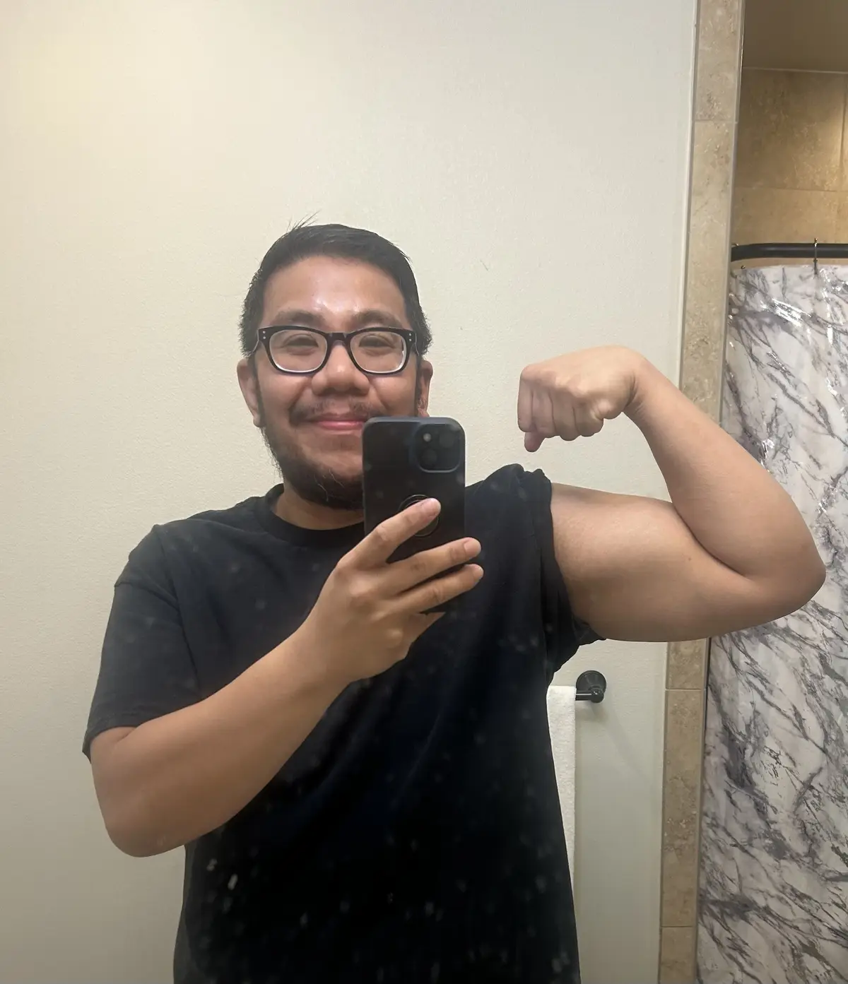 A man with glasses and a beard takes a mirror selfie in a bathroom, flexing his bicep while holding a smartphone. He is…
