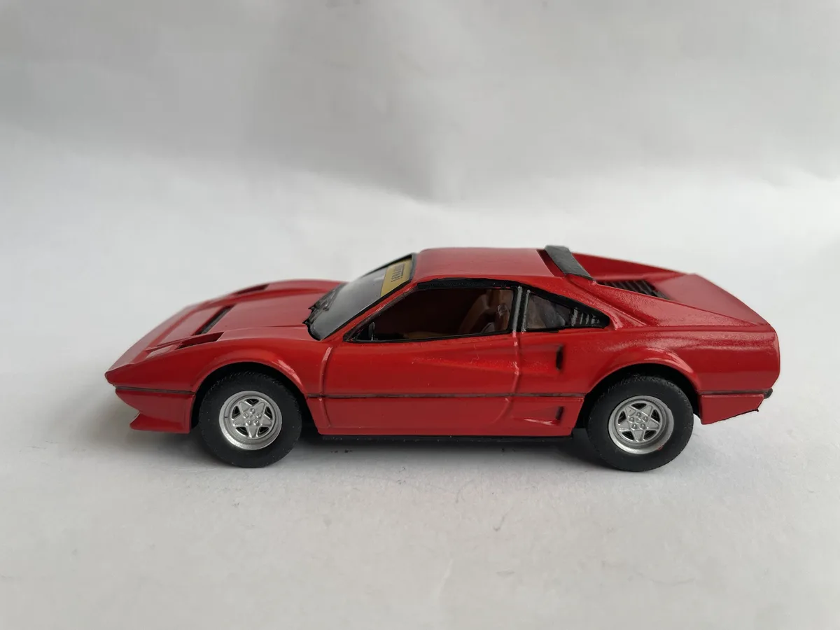 A red toy car, resembling a Ferrari, is shown in profile against a plain white background. The car has silver wheels, black…