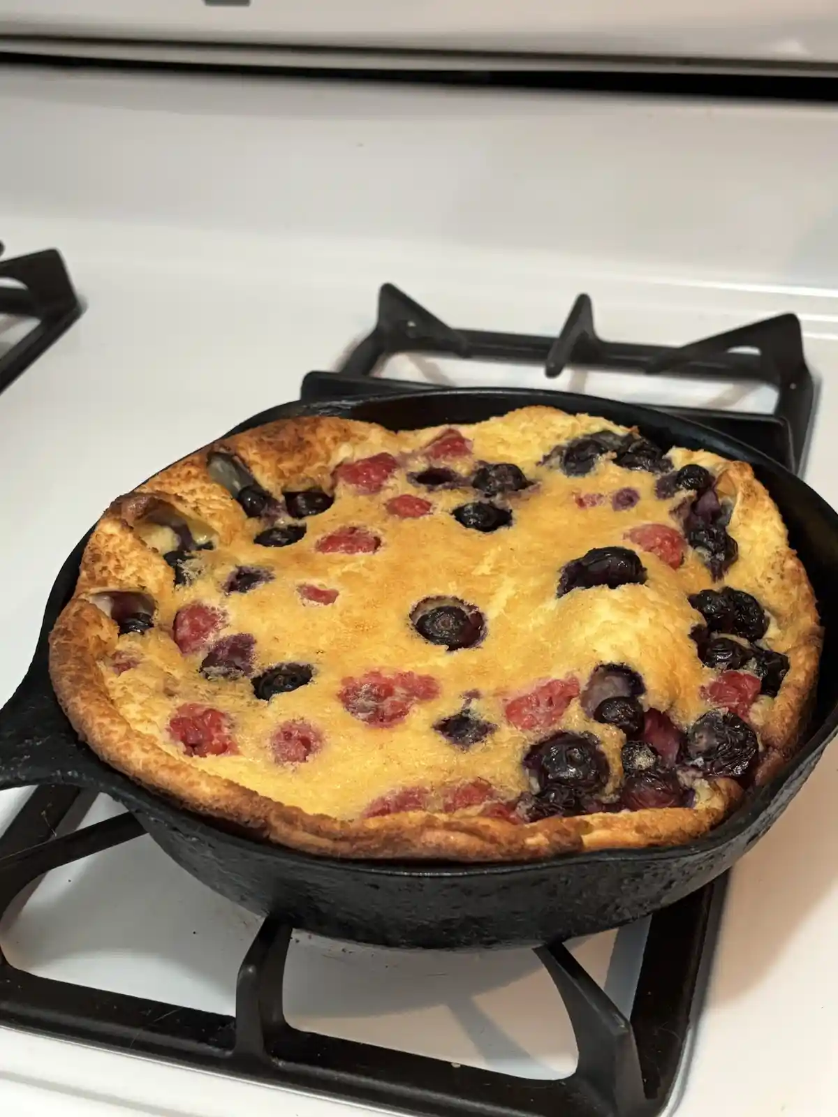 A golden-brown Dutch baby pancake, studded with red raspberries and dark blueberries, sits puffed up in a black cast iron…