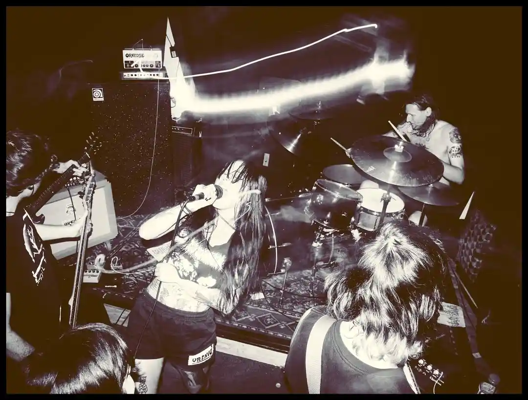 A black-and-white photo of a live rock band performing on stage with a singer, guitarist, and drummer visible. The image has…