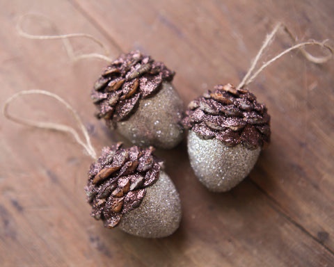 Three glittery acorn ornaments with sparkly silver bodies and coppery-brown caps sit on a wooden surface. Each has a twine…