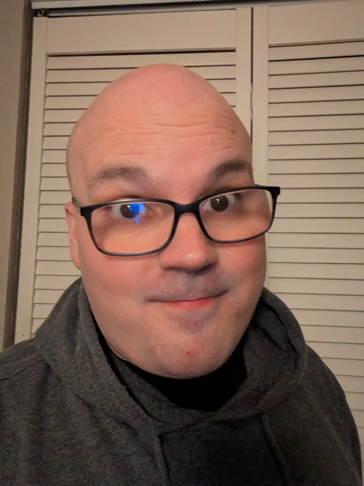 A bald man wearing black-rimmed glasses and a grey hoodie stares directly at the camera with wide eyes and a slight smirk…