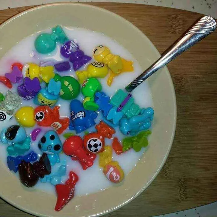 A close-up photo shows a white bowl filled with milk and numerous colorful plastic toys, resembling cereal pieces, with a…