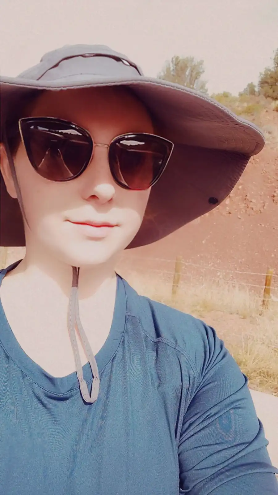 A person wearing a wide-brimmed hat and sunglasses takes a selfie outdoors in a dry, sunlit landscape with a fence and…