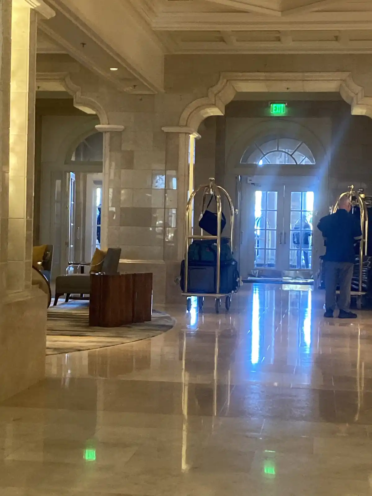 A grand hotel lobby with marble floors, arched doorways, and a green exit sign above a glass door. A bellhop pushes a…