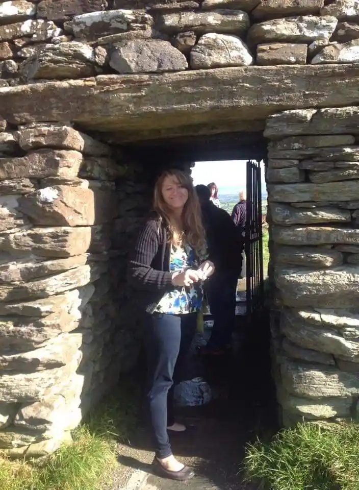 A woman with long blonde hair, wearing a patterned top and dark pants, stands smiling in the narrow stone doorway of an…