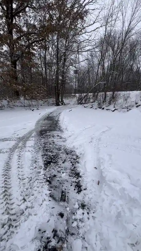 A snowy forest path shows a narrow, icy, and muddy trail flanked by deep snowdrifts and bare trees under an overcast sky…