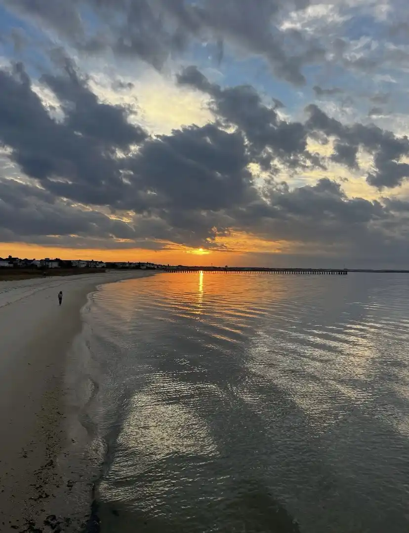 A serene beach sunset with dramatic clouds, a long pier extending into the water, and a lone person walking along the…