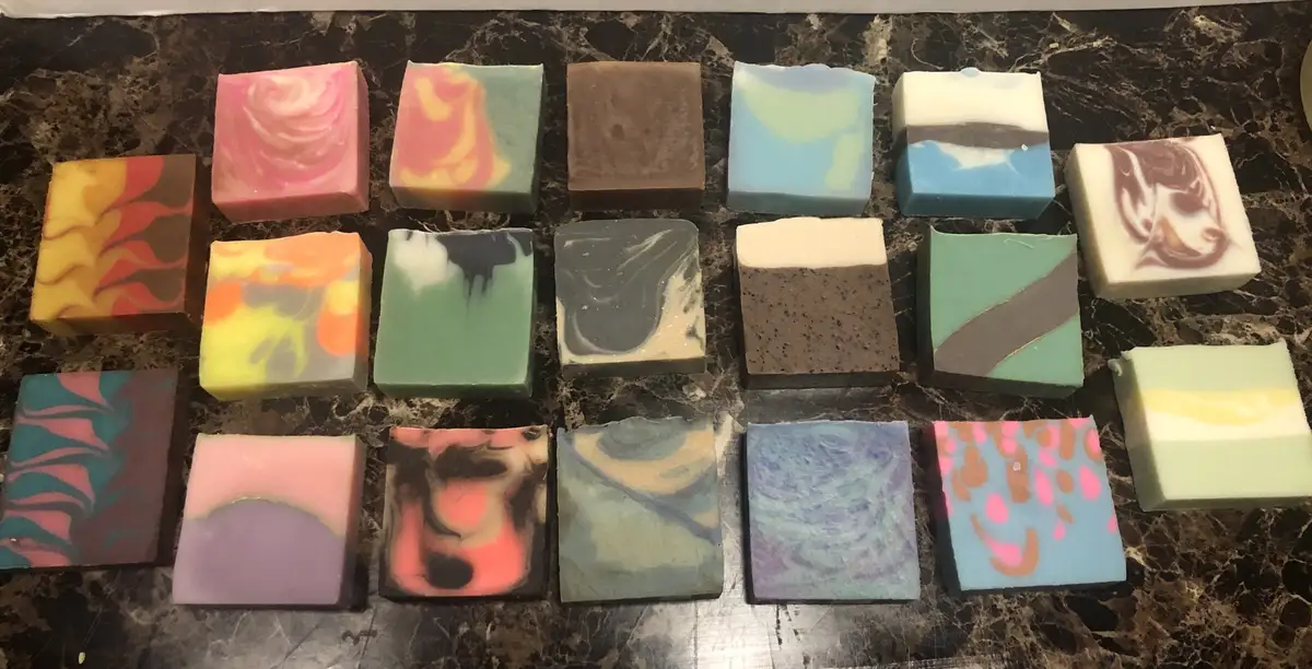 An overhead shot displays a collection of 16 handmade, square-shaped soaps with vibrant, swirling, and layered colors…