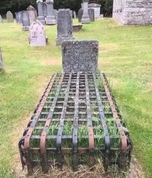 A weathered, moss-covered gravestone in a cemetery is humorously covered by a rusty, metal grid resembling a bed frame, with…
