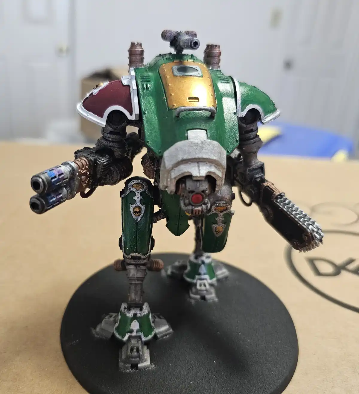 A heavily armored, green and gold painted war machine miniature stands on a black circular base, holding weapons in both…