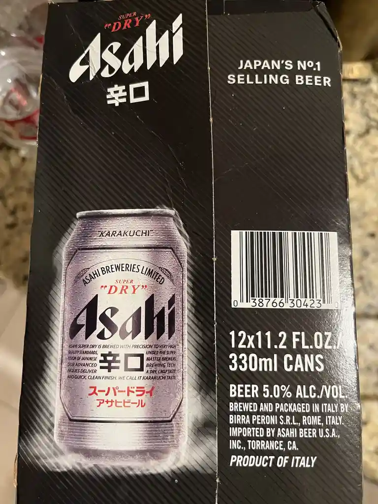 A close-up of an Asahi Super Dry beer cardboard box, showing its black packaging with white and red text. The front displays…