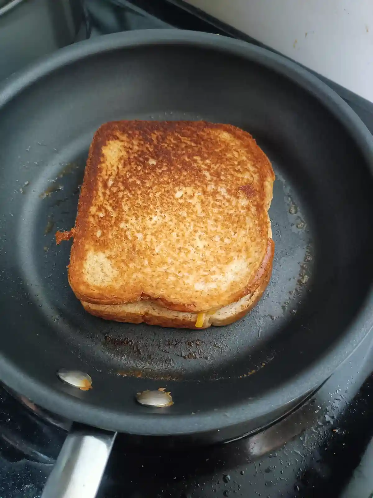 A close-up photo shows a perfectly golden-brown grilled cheese sandwich sizzling in a black non-stick frying pan on a…