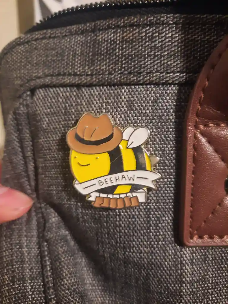 A close-up photo shows a cartoon bee pin attached to a textured gray fabric, likely a bag or jacket. The bee is wearing a…